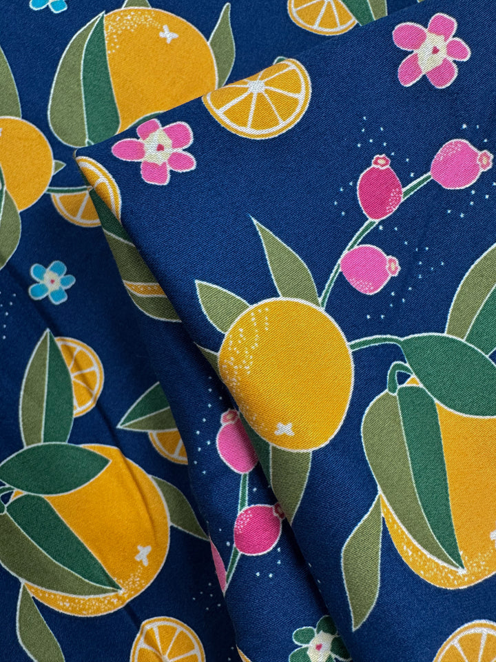 Cotton Sateen - OJ by Super Cheap Fabrics is a medium weight cotton fabric (145cm wide) with a vibrant print of yellow oranges, orange slices, pink flowers, and green leaves on a dark blue background. Pattern repeats are visible on the folded fabric.