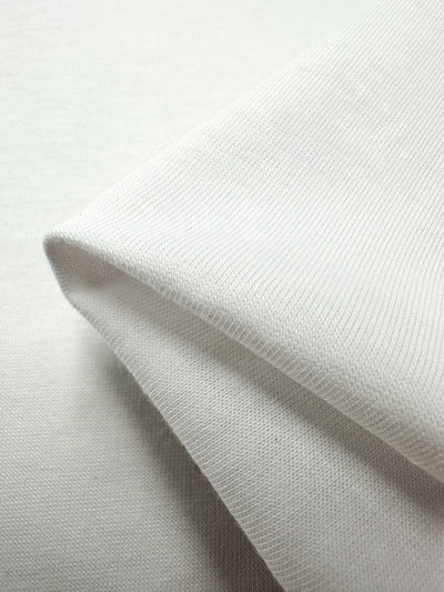 Close-up of a folded piece of light white knitted cotton jersey fabric from Super Cheap Fabrics, showcasing its texture and softness. This cotton blend has a smooth and evenly woven appearance, highlighting the details of individual threads and stitches, making it perfect for crafting comfortable dresses.