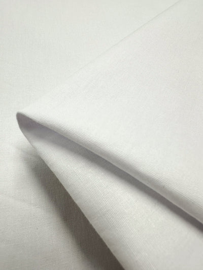 Close-up of a neatly folded piece of Stretch Cotton - White - 125cm from Super Cheap Fabrics, showing its fine texture and smooth surface. The folds are crisp, highlighting its clean and fresh appearance. The background is also white, blending seamlessly with the lightweight fabric's 125 CM width.