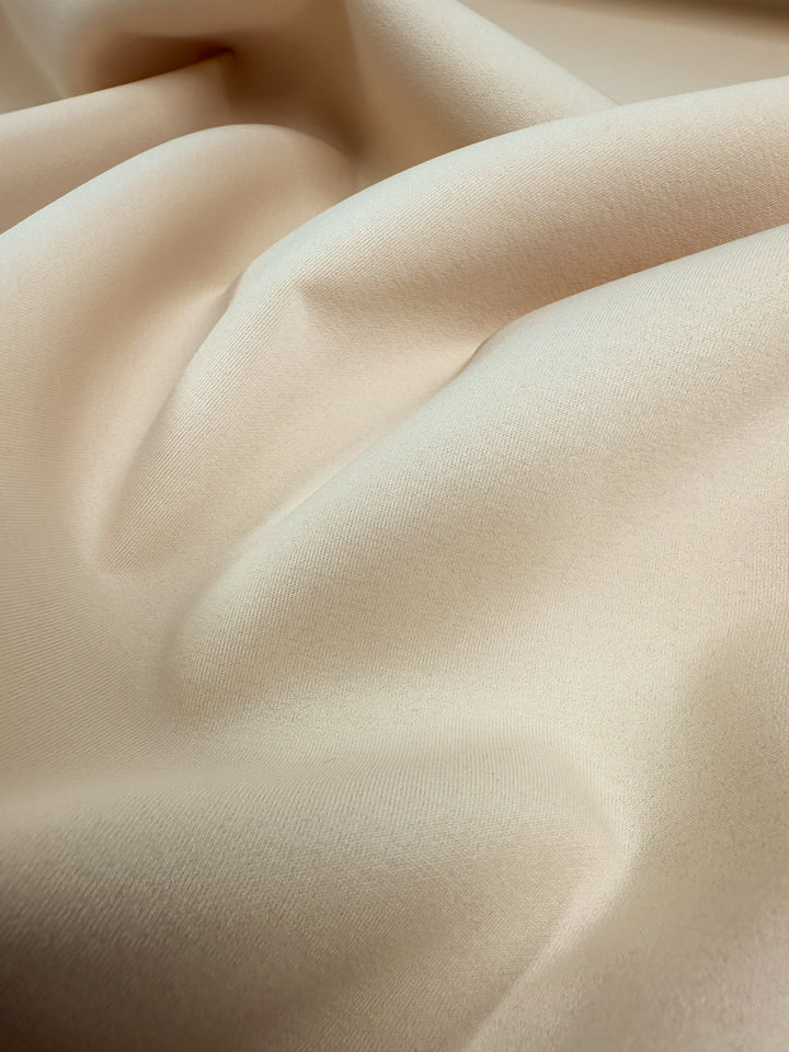 Close-up of Super Cheap Fabrics' Scuba Knit - Mother of Pearl, a smooth, light beige fabric measuring 165cm wide. It features gentle folds and soft creases with a silky, luxurious texture that reflects a subtle sheen under the light.