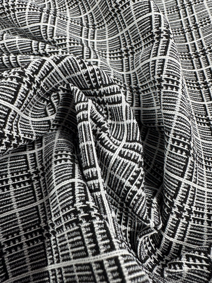 Close-up of crumpled medium-weight Textured Bengaline - Plaidly fabric from Super Cheap Fabrics. The detailed black and white geometric pattern, featuring intersecting lines that form various shapes, creates an intricate and textured appearance. This 150cm wide multi-use fabric appears soft and silky.