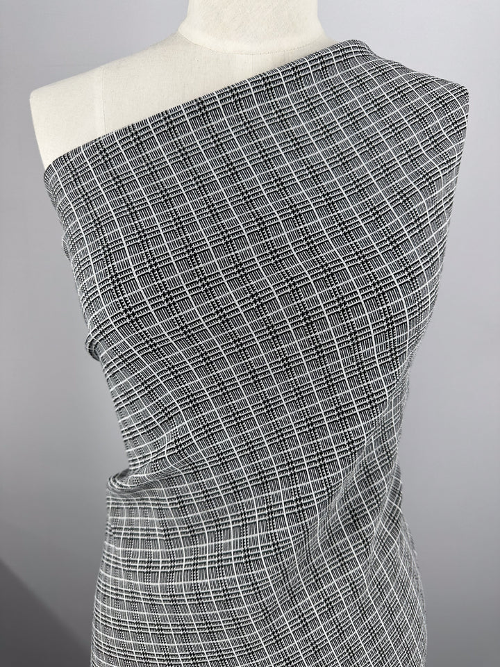 A mannequin draped in Super Cheap Fabrics' Textured Bengaline - Plaidly - 150cm. The medium-weight fabric showcases a delicate, overlapping grid pattern and is wrapped asymmetrically, covering one shoulder while leaving the other exposed. In the background is a plain, light gray backdrop.