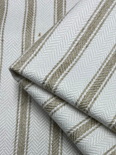 A close-up image of folded Upholstery Herringbone - Salt & Sand - 145cm by Super Cheap Fabrics featuring a herringbone weave pattern. The fabric is primarily white, accented with evenly spaced vertical beige stripes. The texture of the weave and the stripes are clearly visible, showcasing its durable and stylish qualities in an affordable range.