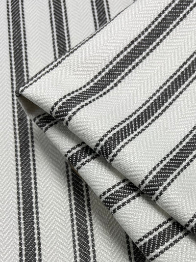 Close-up image of a folded Upholstery Herringbone - Tea Leaf - 145cm by Super Cheap Fabrics featuring a white background with parallel black and gray stripes. The texture of the fabric showcases a subtle chevron pattern that adds to the material's detail and depth, making it part of an affordable range of durable fabrics.