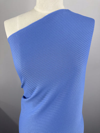 A close-up of a mannequin draped in Super Cheap Fabrics' Textured Knit - Ultramarine - 155cm. The medium weight, light blue polyester fabric features a unique honeycomb-like pattern and is elegantly wrapped over one shoulder, creating an asymmetrical look against a neutral gray background.