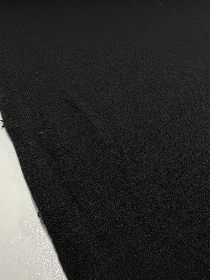 A close-up photo of the Textured Knit - Black - 150cm fabric by Super Cheap Fabrics, showcasing its medium weight and smooth texture, with minor imperfections. Ideal for dresses, this fabric appears to be draped or laid out on a flat surface. The lighting highlights the subtle details in the weave, bringing out its texture and sheen.