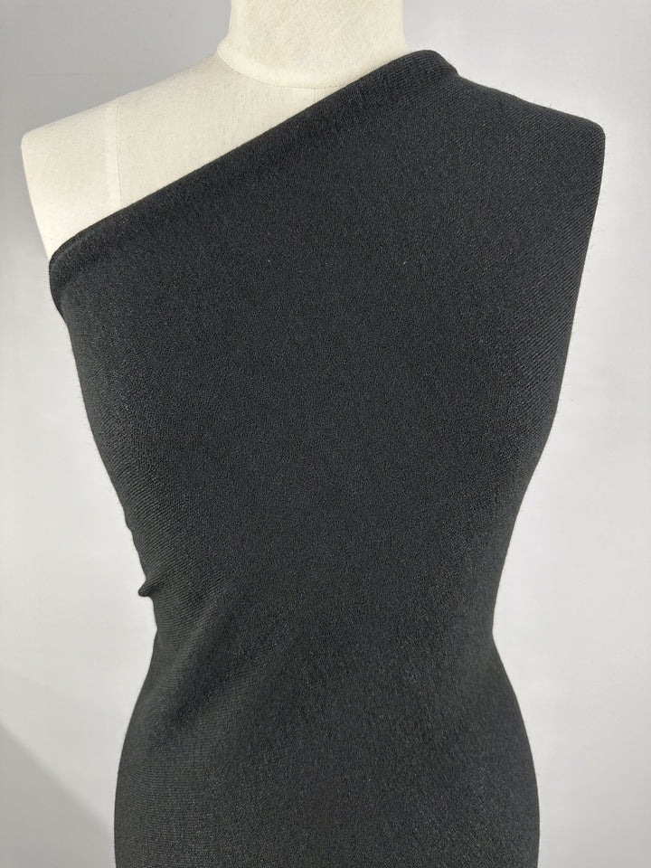 A mannequin is dressed in a one-shoulder, body-hugging dress made from Super Cheap Fabrics' Textured Knit in black. The medium weight fabric, at 150cm wide, drapes elegantly over the form, showcasing a minimalist and chic style against a neutral background.