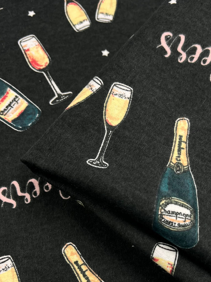 A lightweight fabric with a pattern of champagne bottles and glasses filled with champagne, labeled "champagne sweet wine." The dark background features small star accents, and the image shows a folded section. This multi-colour design is crafted from 100% cotton. This is the Flannelette - Cheers - 110cm by Super Cheap Fabrics.