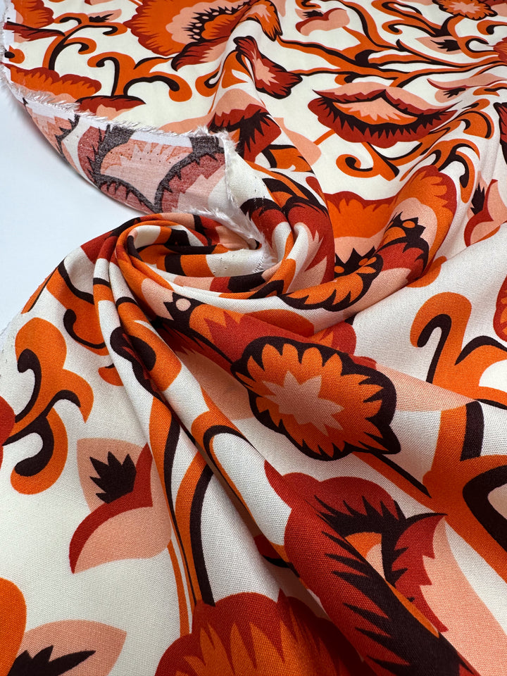 A close-up of Super Cheap Fabrics' Designer Rayon - Floral Delight - 145cm, featuring a swirling floral pattern in orange, red, and cream. This retro-inspired fabric is ideal for versatile clothing and eye-catching prints.