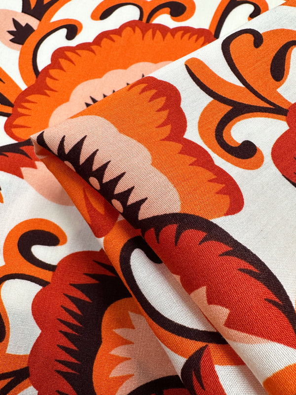 Close-up of Super Cheap Fabrics’ Designer Rayon - Floral Delight - 145cm: rayon fabric with a bold orange, red, and black floral print on cream. Slight folds highlight its intricate, stylized design—ideal for versatile clothing projects.
