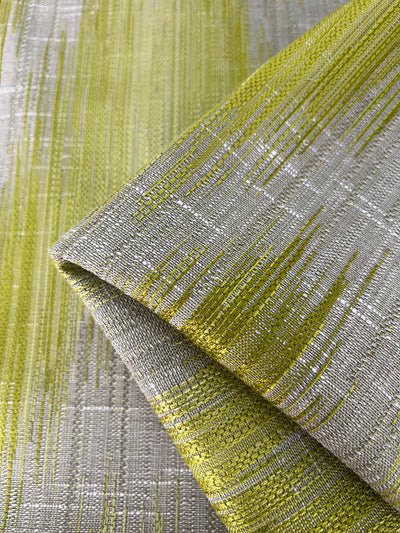 A close-up photo of Upholstery Jacquard - Willow - 145cm from Super Cheap Fabrics featuring a woven design with a green and silver gradient, presenting an abstract pattern. The fabric is partially folded, revealing its detailed texture and the interplay of colors, exemplifying the quality found in durable fabrics from our affordable range.