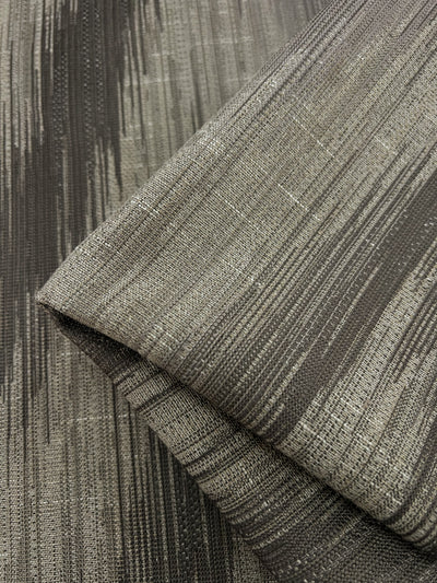 Close-up of a folded piece of fabric with a textured, vertical striped pattern. The Super Cheap Fabrics Upholstery Jacquard - Walnut - 145cm features various shades of gray, giving it a modern and sophisticated appearance. This durable fabric's intricate weave creates a sense of depth and dimension.