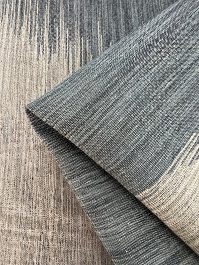 Close-up image of two overlapping pieces of upholstery fabric with varying shades of gray and beige. The fabric has a subtle, linear, textured pattern, creating a gradient effect. The top piece is folded, revealing both sides and highlighting the depth and durability of the Upholstery Jacquard - Peacock - 145cm from Super Cheap Fabrics' affordable range.