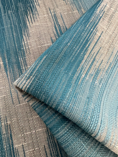Close-up image of textured Upholstery Jacquard - Aqua featuring a blend of teal and beige colors with a subtle wavy pattern. The fabric is folded, showcasing its intricate weaving and pattern from different angles, highlighting the qualities of durable fabrics within an affordable range by Super Cheap Fabrics.