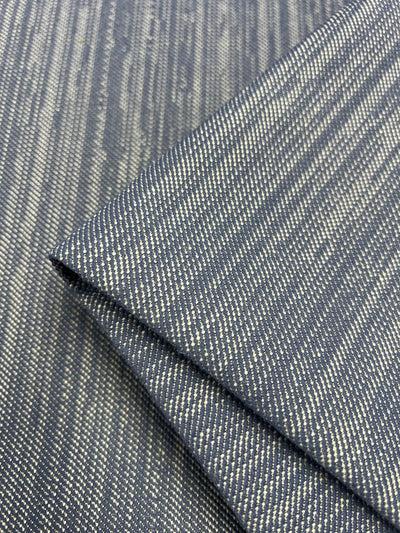 A close-up image of folded fabric with a striped pattern. The fabric is navy blue with thin, light-colored horizontal stripes, creating a textured appearance. The folds emphasize the durable fabric's design and texture, making it ideal for furniture upholstery. This is the Upholstery Twill - Lavendar Blue - 147cm by Super Cheap Fabrics.