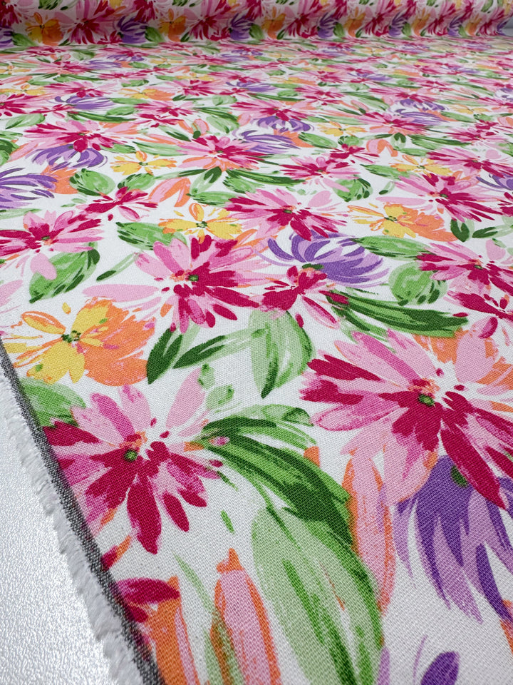 A close-up of Super Cheap Fabrics’ Printed Linen Blend - Petal Parade (135cm) shows a vibrant floral pattern in pink, purple, yellow, and orange with green leaves on white—ideal for summer jackets or upholstery.