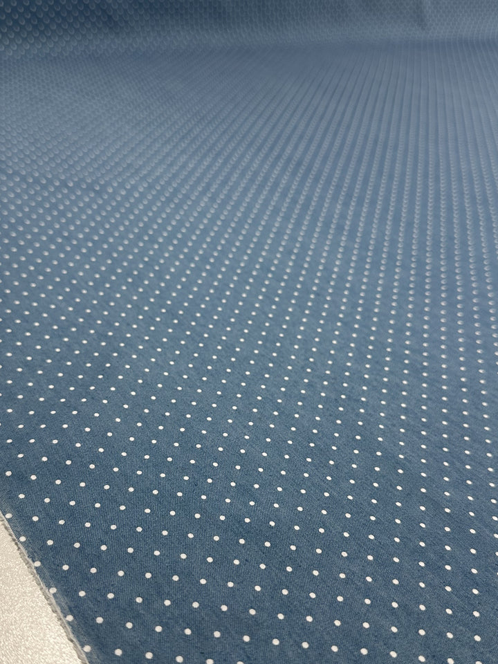A close-up of Super Cheap Fabrics' Chambray - Blissful Spot - 150cm shows its lightweight blue fabric with evenly spaced small white polka dots, creating a textured, visually repetitive surface.