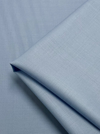 A close-up shot of neatly folded light blue fabric with luxurious texture. The smooth fabric, featuring a subtle weave pattern, boasts a clean and polished appearance. The image focuses on the folds and the material's fine details, revealing its moisture-wicking properties. This is the Merino Wool Suiting - Serenity - 155cm by Super Cheap Fabrics.