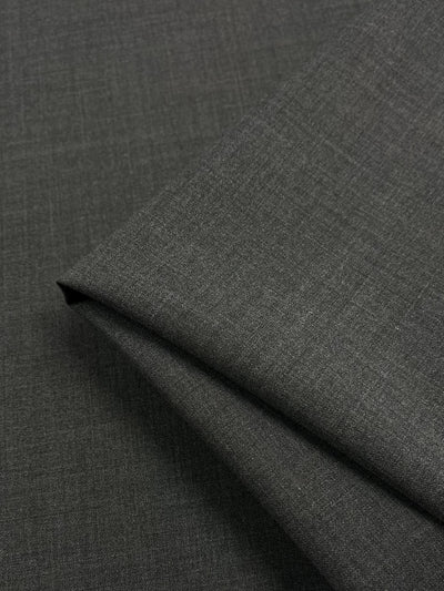 A close-up view of a folded piece of dark gray fabric. The Super Cheap Fabrics Wool Suiting - Charcoal - 152cm has a subtle, luxurious texture, giving it a slightly heathered appearance. The folds in the fabric create gentle, angular lines, highlighting its moisture-wicking properties.