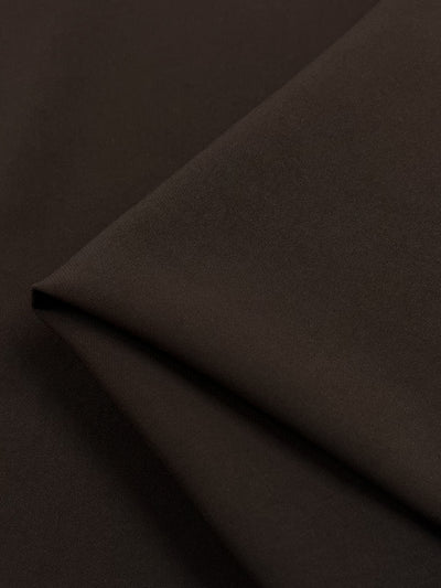 A close-up image of folded dark brown Premium Viscose Suiting - Choc Martini - 130cm from Super Cheap Fabrics. The material appears to have a smooth texture and is neatly arranged with visible folds creating an angular pattern. The lighting emphasizes the rich, deep brown color reminiscent of Choc Martini and highlights the subtle sheen of this medium weight fabric.