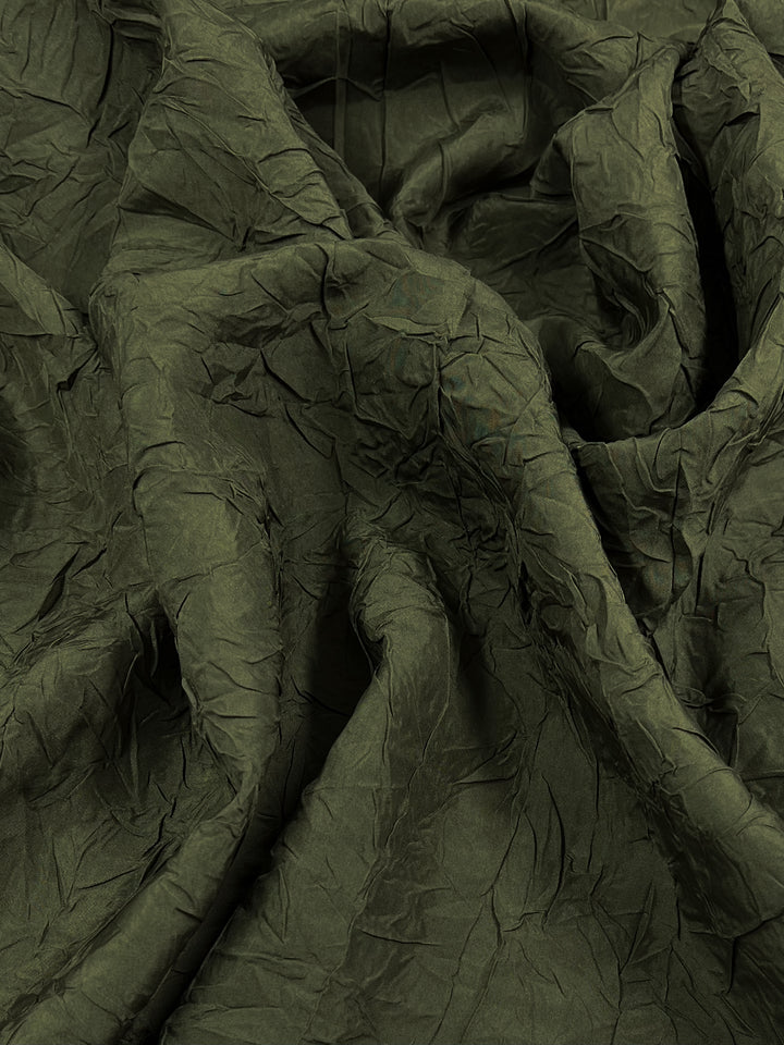 A close-up view of crumpled, dark green Worsted Crinkle Wool - Olive - 125cm from Super Cheap Fabrics with visible texture and folds. The material appears to be lightweight and has an uneven, wrinkled surface. The shadows and highlights emphasize the intricate creases throughout the high-quality wool fibers.