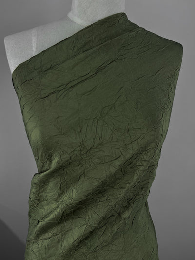 A close-up image of a mannequin draped in luxurious, olive green Worsted Crinkle Wool - Olive - 125cm by Super Cheap Fabrics. The high-quality wool fibers are wrapped around the mannequin from the left shoulder across to the right side, creating an asymmetrical look. The background is a solid gray.