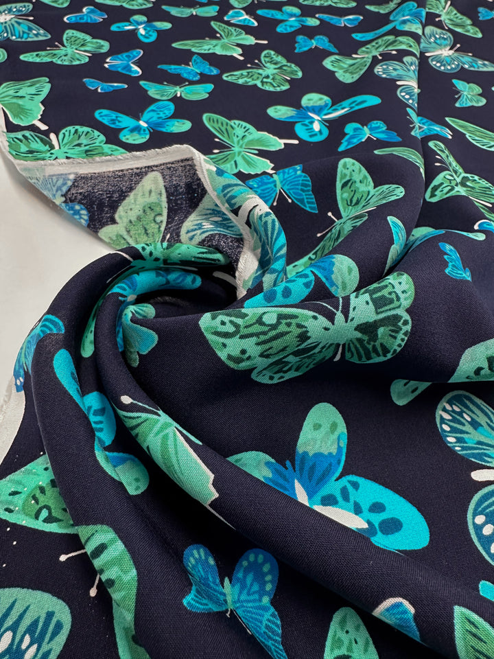 A close-up of Super Cheap Fabrics’ Designer Rayon - Midnight Flutter (145cm) reveals a vibrant butterfly pattern in blue, teal, and green. The partially rolled fabric displays both the front and back sides of its striking prints.