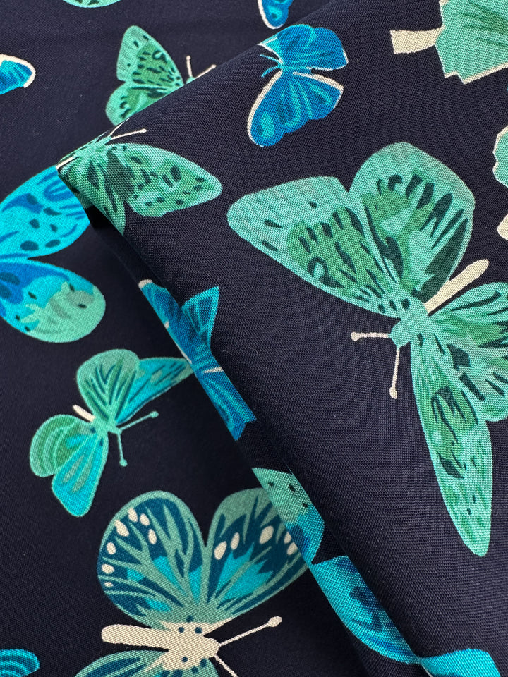 Designer Rayon - Midnight Flutter by Super Cheap Fabrics is a 145cm wide dark rayon fabric, featuring vibrant green and blue butterfly prints with white accents, overlapping in various orientations.