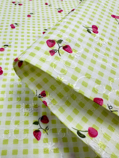 Here's a close-up of the Embroidered Printed Cotton - Strawberry Lime fabric by Super Cheap Fabrics. This 114cm wide material highlights green checkered patterns adorned with pink strawberries. Made from natural cotton fibers, it is expertly folded to reveal delicate white embroidery outlining the strawberries. With its vibrant and joyful design, this fabric is ideal for creating clothing and household items.