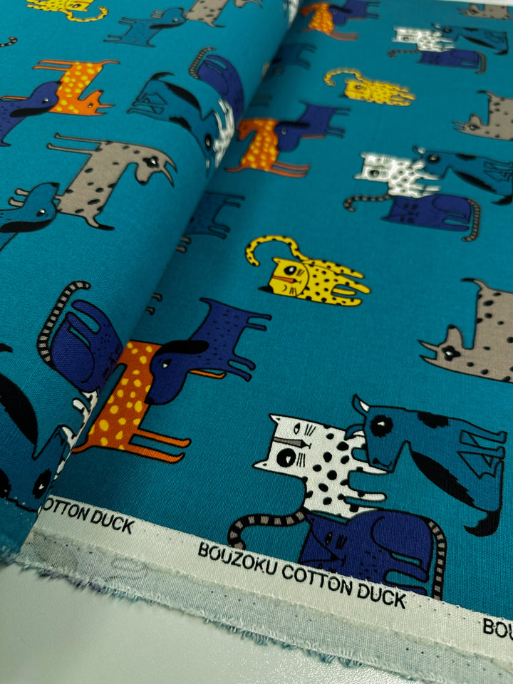 Fabric showcasing charming cat illustrations in a variety of colors and designs, such as polka dots and stripes, set against a teal background. This medium-weight Cotton Duck fabric is ideal for home décor projects. The edge of the fabric reads "Cotton Duck - Cat Dog - 112cm." It is offered by Super Cheap Fabrics.