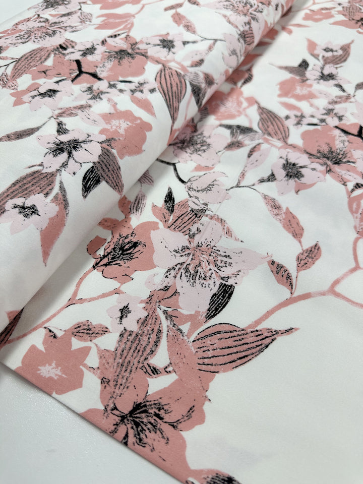 A close-up of the Cotton Sateen - Lizzy's Garden by Super Cheap Fabrics, highlighting its floral pattern with pink and black flowers set against a white backdrop. This lightweight fabric is ideal for household décor items, offering intricate and detailed designs in a range of shades, providing a delicate appearance.