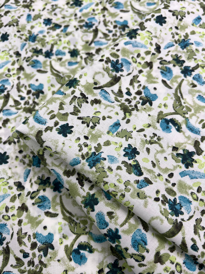 Close-up of a folded piece of Super Cheap Fabrics' "Printed Cotton - Blue Natures Mark - 148cm," featuring a vibrant floral pattern with blue, green, and black flowers scattered throughout. This lightweight printed cotton fabric displays a smooth texture with intricate details in both the flowers and leaves.