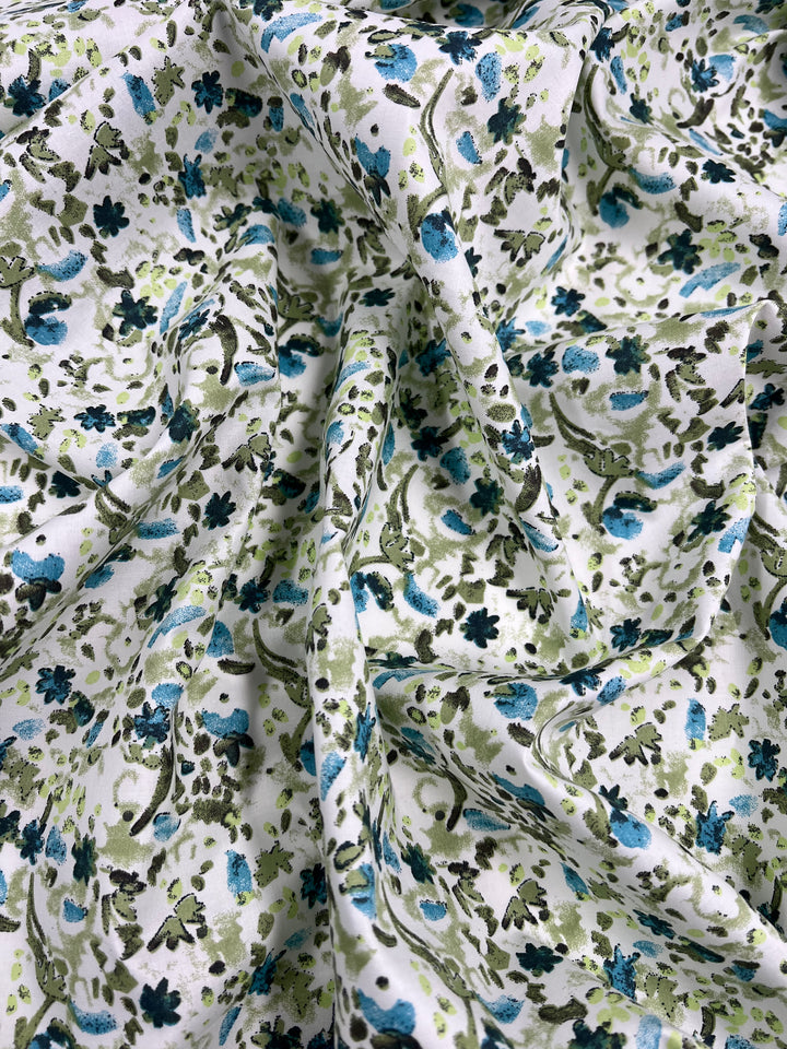 A close-up image of the Super Cheap Fabrics' Printed Cotton - Blue Natures Mark - 148cm showcases its 100% cotton fabric with a white background decorated with small flowers and leaves in shades of blue, green, and black. The lightweight material appears to be soft and slightly crumpled.