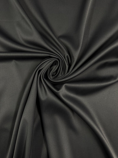 Super Cheap Fabrics' Satin Deluxe - Forged Iron (140cm) features smooth black satin with spiral folds and a swirling texture, highlighting its shiny finish.