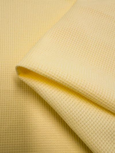 Close-up of a folded, yellow Waffle Knit - Pale Banana - 170cm from Super Cheap Fabrics. The Waffle Knit - Pale Banana's texture, resembling honeycomb fabric, includes small, repeating grid patterns characteristic of waffle-weave fabric. The image highlights the three-dimensional effect and soft, cozy material.