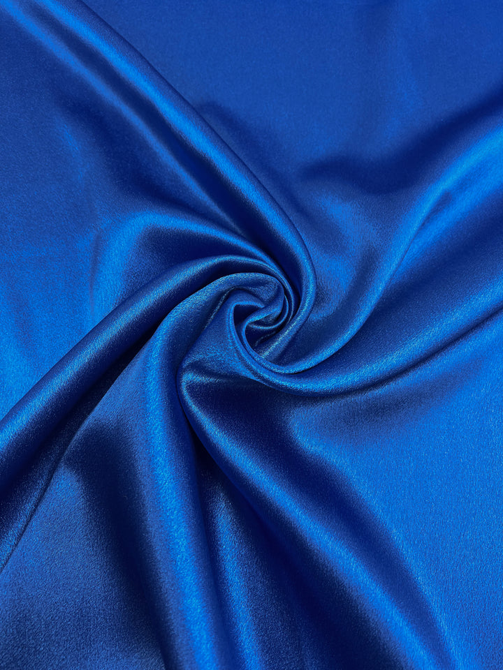 A close-up view of the Satin Back Crepe - Royal - 150cm by Super Cheap Fabrics showcases its shiny blue surface with a swirling fold in the center, highlighting its smooth and reflective texture, ideal for evening and casual wear.