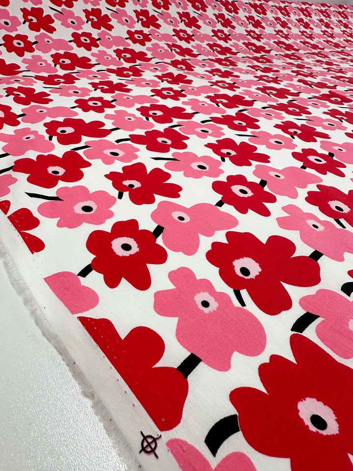 A dynamic piece of "Little Miss Sunshine" printed cotton fabric from Super Cheap Fabrics, displaying a striking pattern of red and pink flowers on a white backdrop with prominent black stems and centers, ideal for various applications.