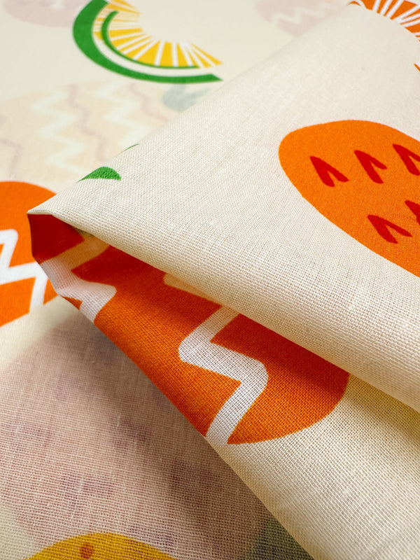 A detailed view of the Super Cheap Fabrics' Printed Cotton - Summer displays a lively assortment of abstract fruit patterns. The design includes bright orange, green, and yellow fruit shapes with zigzag motifs set against a cream backdrop. This lightweight fabric is neatly folded, highlighting its vibrant and playful design.