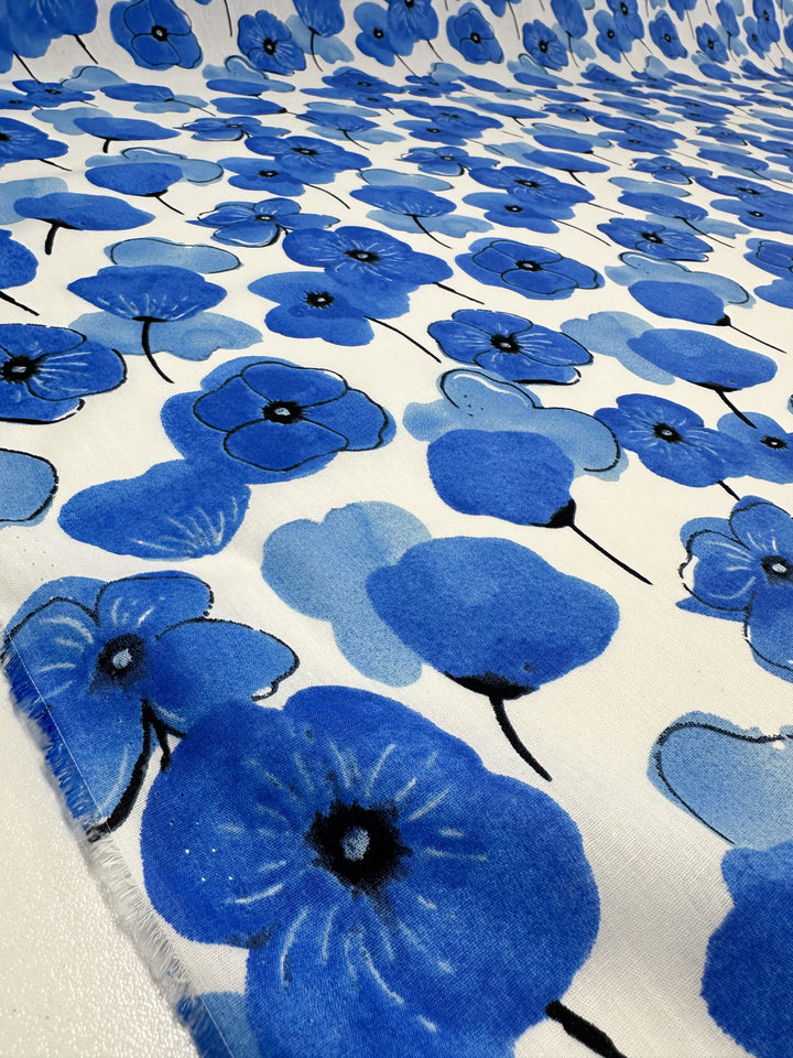 The Printed Cotton - Mykonos - 150cm from Super Cheap Fabrics displays a pattern featuring blue poppies with black centers set against a white background. The seamless design beautifully spans the entire fabric, making it ideal for creating both clothing and household items using natural cotton fibers.