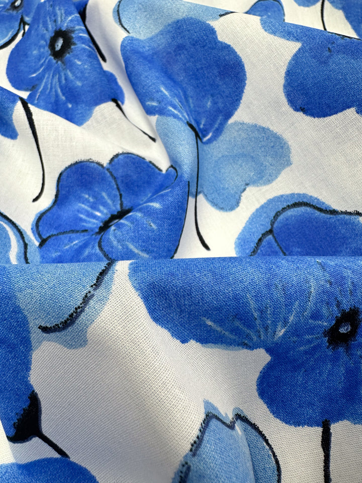 A close-up of Super Cheap Fabrics' Printed Cotton - Mykonos - 150cm showcases a pattern of large, vibrant blue flowers with black accents set against a white background. The flowers, made from natural cotton fibers, vary in size and shade, resulting in a bold and striking design.