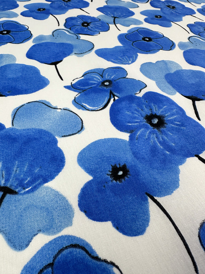 A detailed view of the "Printed Cotton - Mykonos - 150cm" by Super Cheap Fabrics showcases a pattern of blue flowers with black centers set against a white backdrop. Made from natural cotton fibers, the flowers vary in size to create a dynamic and vibrant design.