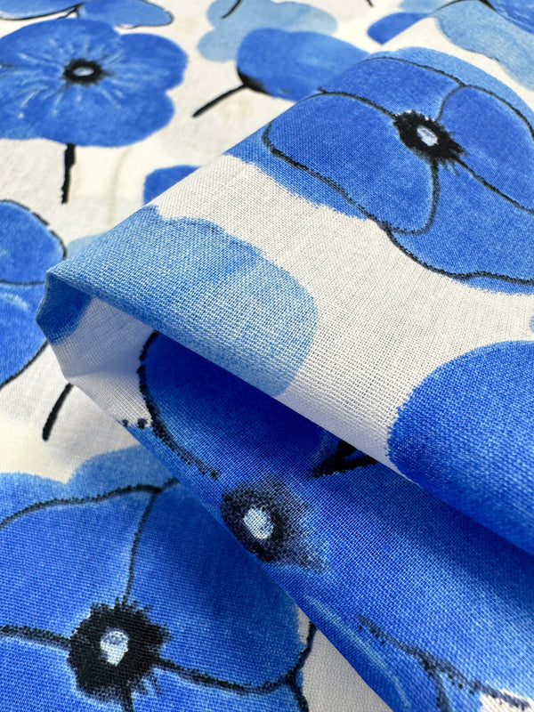 Close-up of Super Cheap Fabrics' Printed Cotton - Mykonos - 150cm, showcasing a vivid floral design. The fabric features large blue flowers against a white background and is made from natural cotton fibers. Its slightly folded state highlights the texture and vibrant pattern.