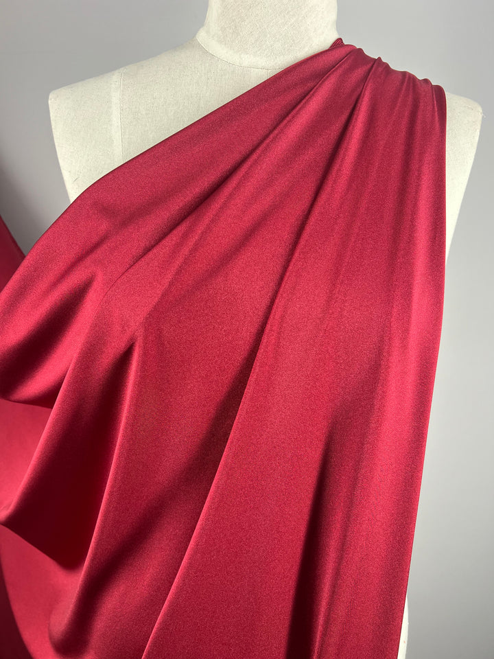 A close-up of a mannequin draped in **Super Cheap Fabrics' Nylon Lycra - Rumba Red - 160cm**. The material is smooth and slightly reflective, showcasing a rich and luxurious texture. The folds of the four-way stretch fabric create soft lines and shadows, emphasizing its fluidity and drape.