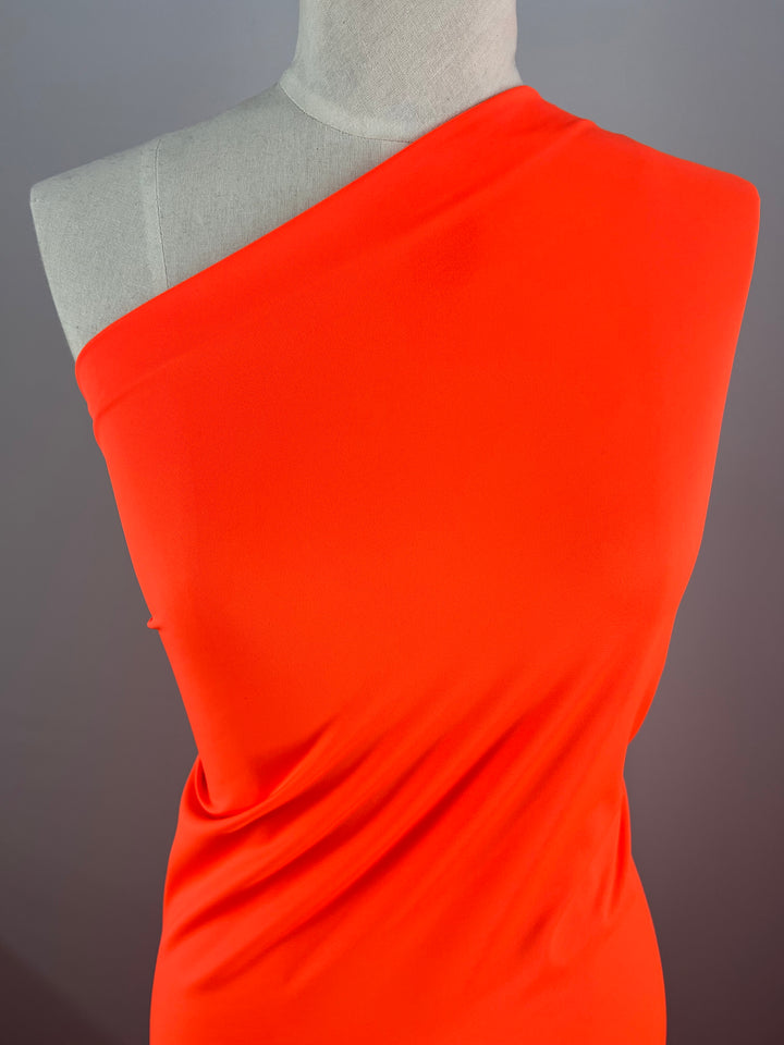 A close-up view of a mannequin dressed in a bright red, one-shoulder garment made from **Super Cheap Fabrics' Nylon Lycra - Fluro Orange - 160cm**. The smooth and form-fitting swimwear fabric showcases a minimalistic and elegant design. The background is a plain gray, putting the focus entirely on the striking red garment.