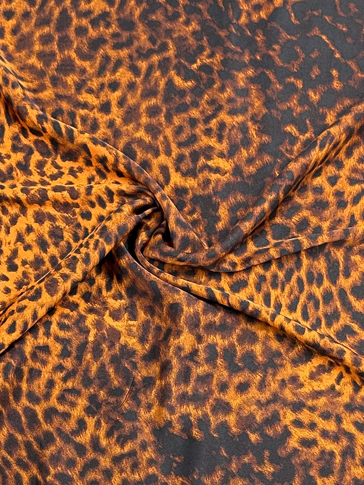 A close-up of Super Cheap Fabrics' Silky Satin - Namer - 145cm, showcasing its leopard print pattern in vibrant shades of orange and black, elegantly draped with a subtle twist at the center.