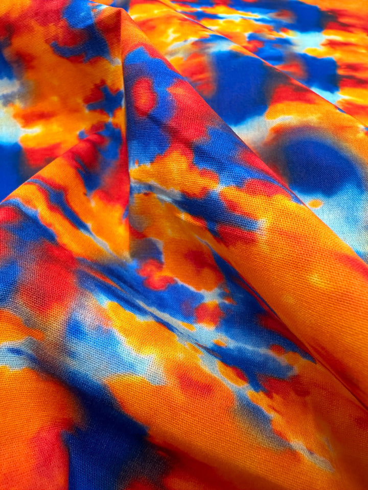 A detailed view of the Printed Cotton - Rosch - 150cm from Super Cheap Fabrics reveals a vibrant abstract pattern. The natural cotton fibers display vivid hues of orange, blue, yellow, and red that blend seamlessly, evoking a dynamic and fiery look with smooth folds and textures.