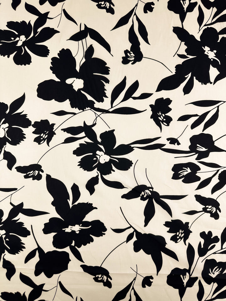 The "Printed Cotton - Visage - 150cm" by Super Cheap Fabrics showcases a stylish black and white floral design with large, stylized flowers and leaves on a light backdrop. This cotton fabric, crafted from natural fibers, offers an artistic arrangement ideal for creating both clothing items and home decor with bold elegance.