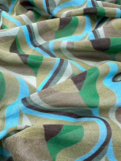 The Pure Printed Silk Voile by Super Cheap Fabrics, named Peacock, displays a wavy pattern in green, blue, and brown. This fabric features smooth, curving lines that form an abstract and fluid design, perfect for creating ultra-lightweight dresses. The material is 140cm wide.