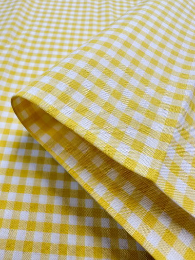 A close-up of the Cotton Gingham Mini - Buttercup from Super Cheap Fabrics showcases its vibrant yellow and white checkered pattern, expertly crafted from natural cotton fibers. The neatly folded fabric displays a geometric grid of squares with a visible texture, giving it a fresh and lively appearance.