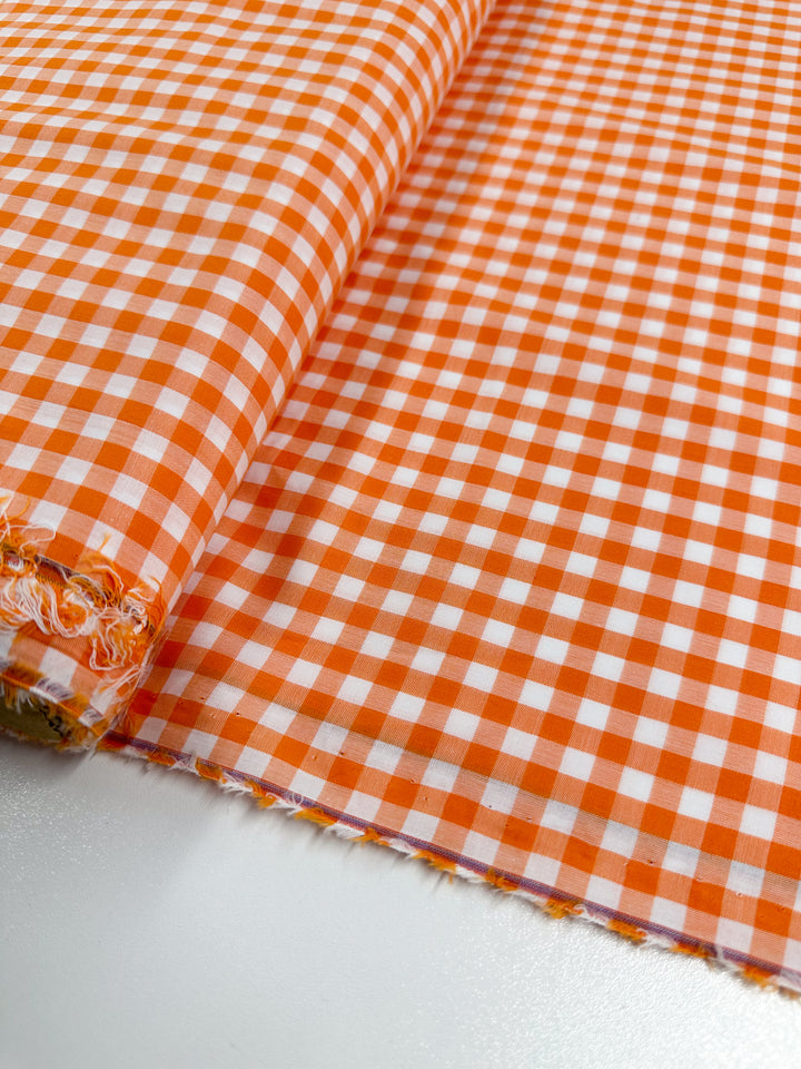 A roll of Cotton Gingham - Mid Orange - 112cm from Super Cheap Fabrics is partially unrolled on a smooth surface, showcasing its natural cotton fibers in an orange and white gingham pattern.